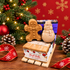 A Christmas-themed gift set featuring a gingerbread man and snowman soap bar stacked on top of a bundle of handmade goat milk soap. Styled on a wooden surface with a red snowflake-patterned backdrop, evergreen tree, ornaments, pinecones, and red ribbon.