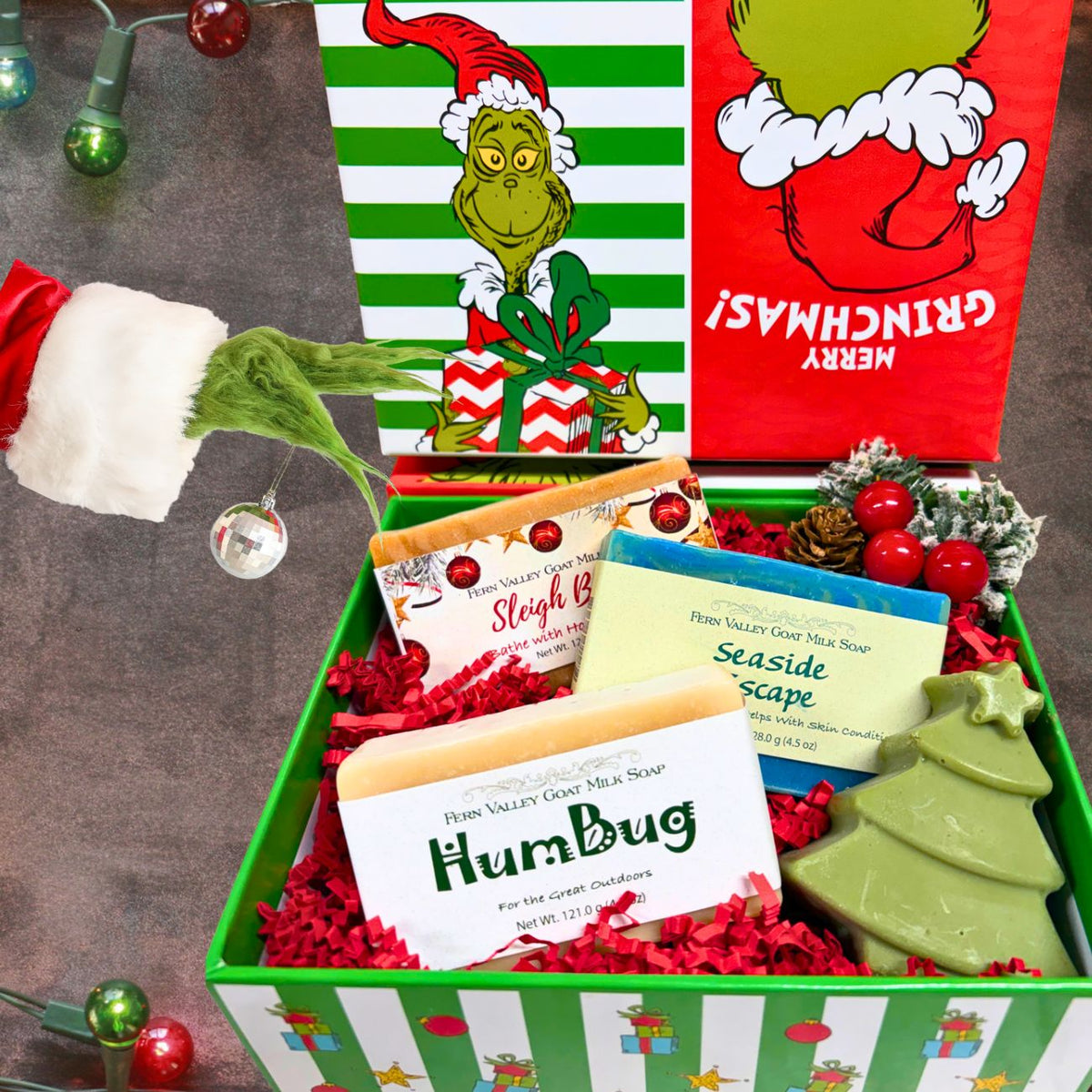 An open green and white striped "Merry Grinchmas!" holiday gift box filled with red crinkle paper, showcasing HumBug, Seaside Escape, and Sleigh Bells goat milk soaps next to a green tree soap, pinecones, and a whimsical green Grinch arm holding a silver ornament.