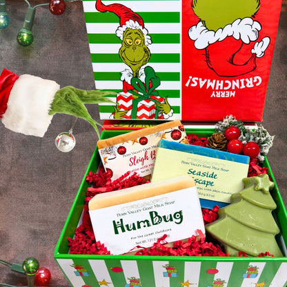 An open green and white striped "Merry Grinchmas!" holiday gift box filled with red crinkle paper, showcasing HumBug, Seaside Escape, and Sleigh Bells goat milk soaps next to a green tree soap, pinecones, and a whimsical green Grinch arm holding a silver ornament.