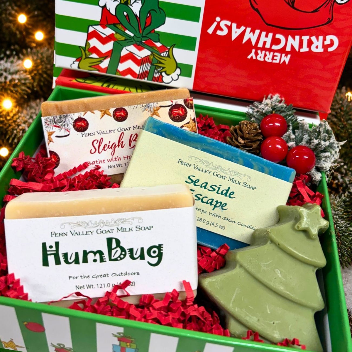 A close-up view of the open Grinch themed gift set displaying three full-size Fern Valley goat milk soap bars labeled HumBug, Seaside Escape, and Sleigh Bells, nestled next to a green Christmas tree soap and bright red winter holiday berries.